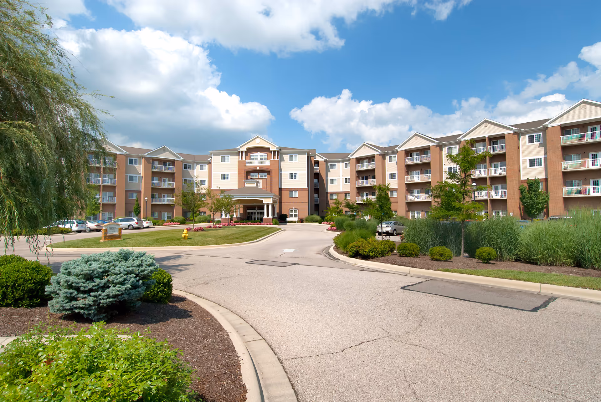 Exterior view of a multi-story senior living facility with a curved driveway, landscaped greenery, and a partly cloudy blue sky.