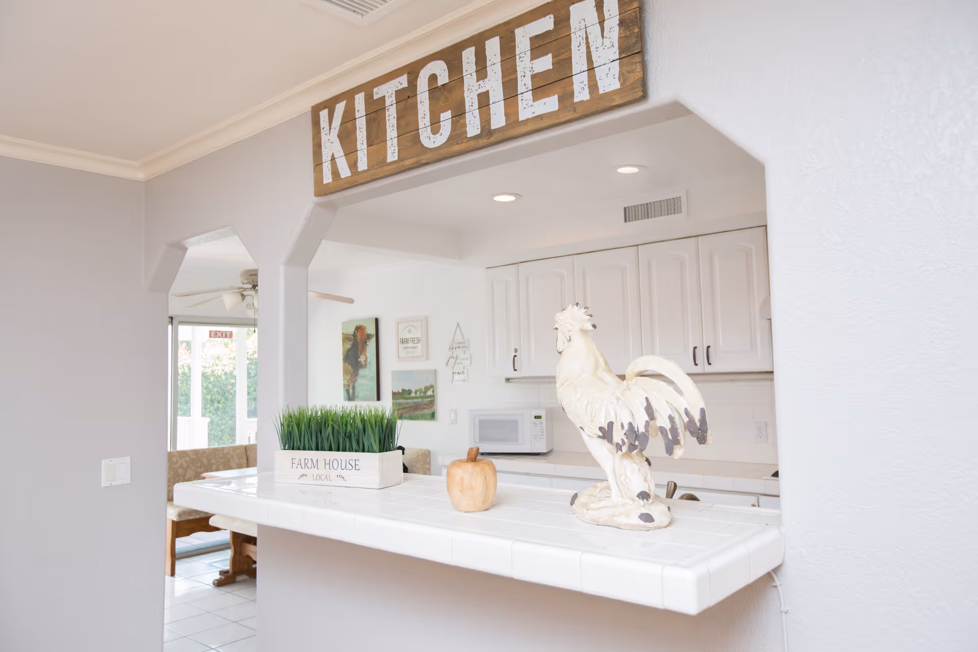 Open kitchen pass-through with a rooster statue on a tiled counter, a planter labeled "FARM HOUSE", and a wooden "KITCHEN" sign above.
