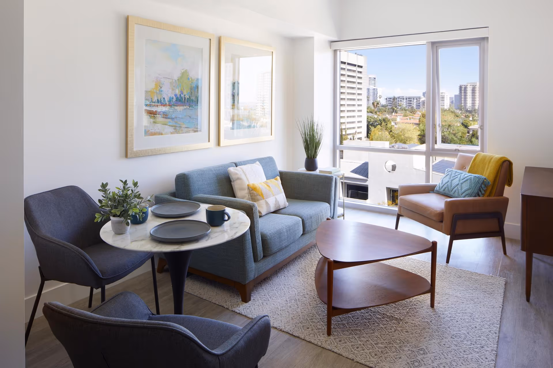 A bright living room with a large window showing a cityscape. The room features a blue loveseat with decorative pillows, a brown armchair with a blue pillow and yellow throw, two dark gray chairs around a small round marble table set with plates and a mug, a wooden triangular coffee table, a light-colored rug, and two framed abstract paintings on the wall.
