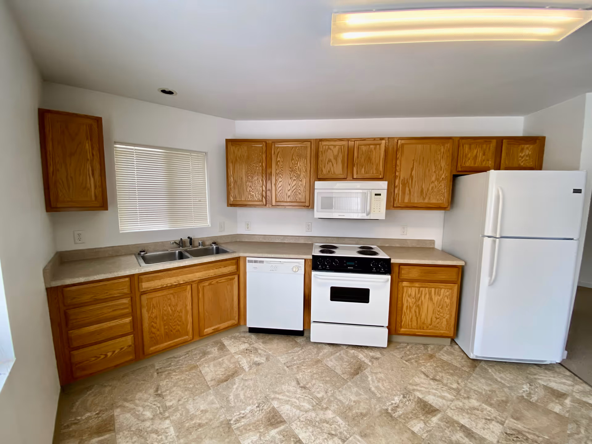 A kitchen with wooden cabinets, a white refrigerator, a white stove with an oven, a white microwave above the stove, a dishwasher, a double sink, and a window with closed blinds. The floor has beige and light brown tiles, and the walls are white.