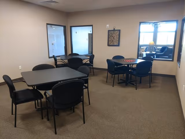 A room with beige walls and carpeted floor containing three black tables, each surrounded by four black chairs. There are two windows on the walls showing another room with seating and a lamp. A decorative clock hangs on the wall between the windows.