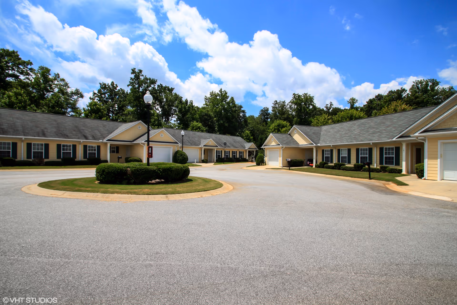 A circular driveway surrounded by single-story residential buildings with garages, beige siding, and dark green shutters under a blue sky with scattered clouds. There are trimmed bushes and a lamppost in the center of the driveway, with trees in the background.