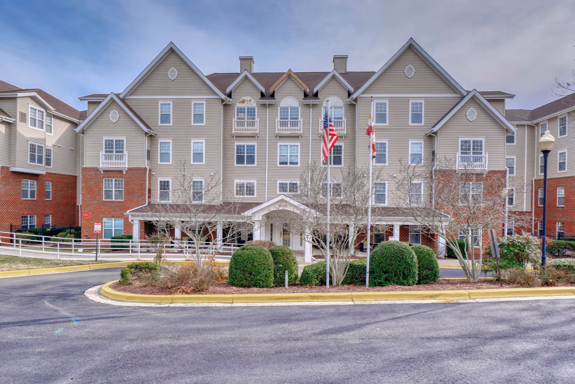 Front exterior of a multi-story senior living building with a covered entrance, flagpoles, and a landscaped circular driveway.