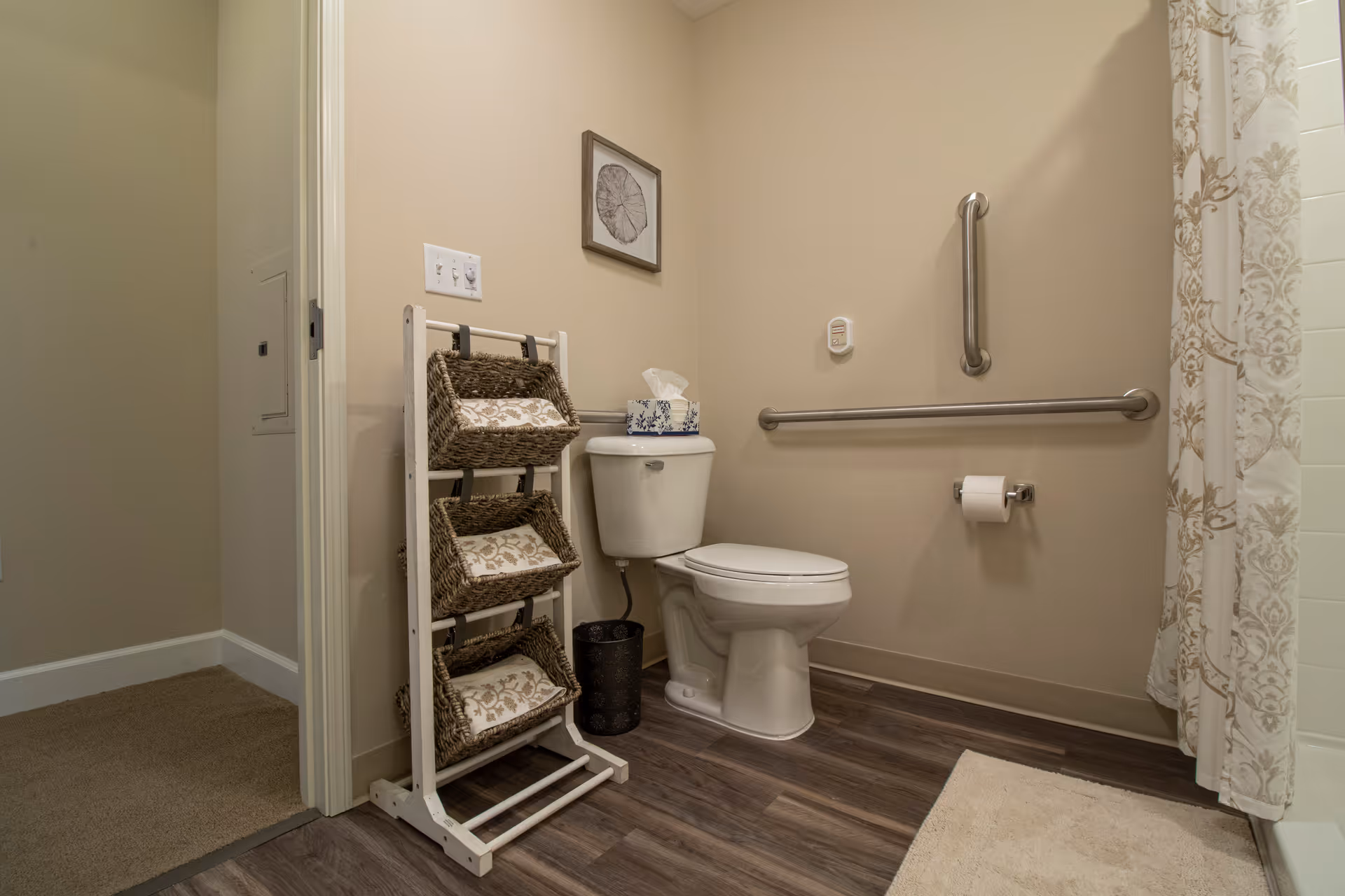 A bathroom with a white toilet, a metal grab bar on the wall, a toilet paper holder with a roll of toilet paper, a three-tier wicker basket stand with patterned towels, a small trash can, a framed artwork on the wall, and a shower curtain with a beige and white pattern. The floor is wood-style vinyl, and the walls are painted beige.
