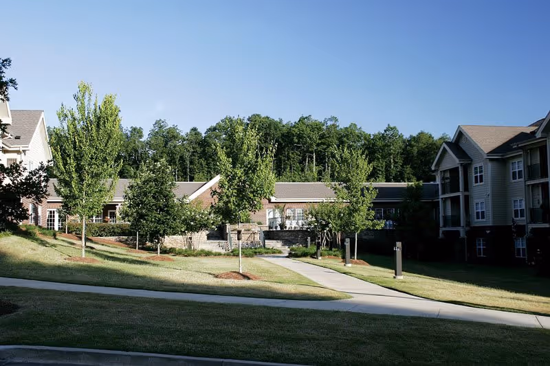 Outdoor view of a senior living facility with a paved walkway, green grass, young trees, and multi-story residential buildings under a clear blue sky.