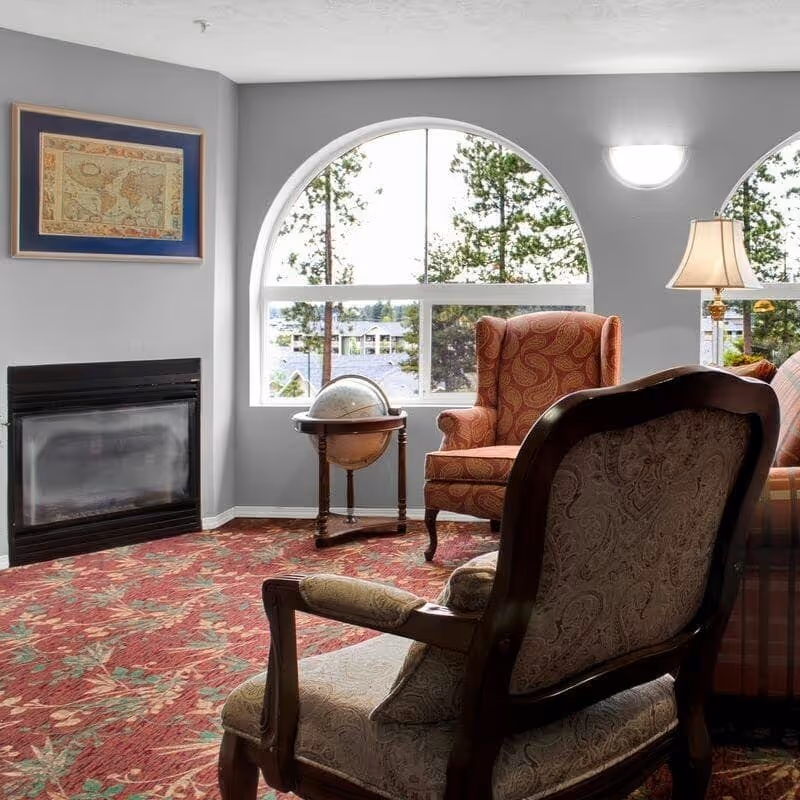 Cozy sitting area with upholstered armchairs around a globe table, a fireplace, and a large arched window showing trees and buildings outside.