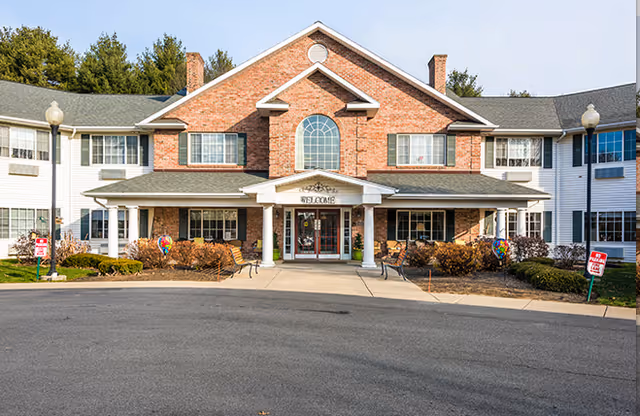 Front facade of The Landing at Queensbury senior living building with a brick central entrance, white columns, landscaping, and a circular driveway.