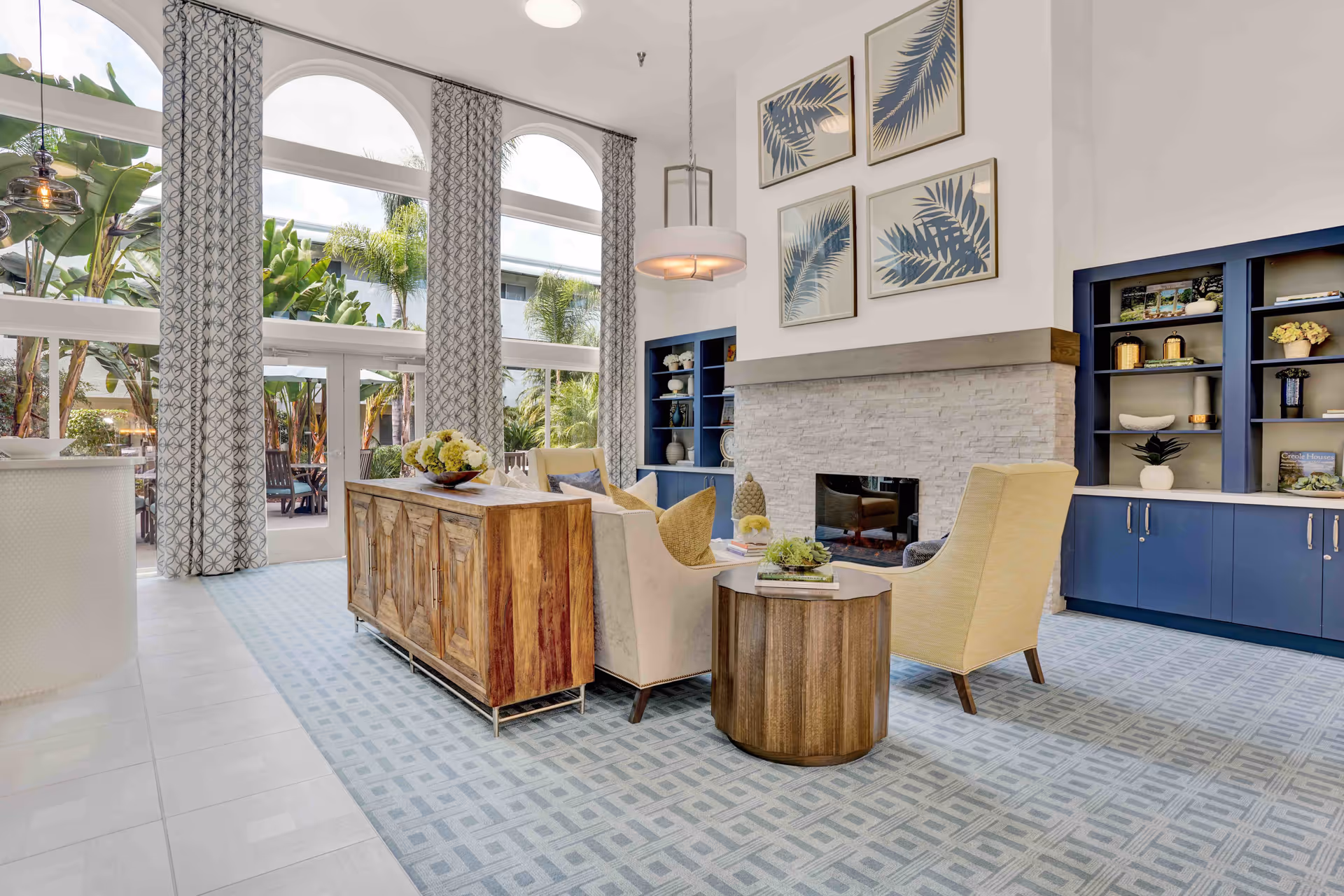 Bright and spacious senior living common area with large arched windows and patterned curtains letting in natural light. The room features a modern fireplace with white stone surround, blue built-in shelves with decorative items, a wooden sideboard, a round wooden side table, and comfortable beige and yellow armchairs arranged on a patterned blue carpet. Outside, lush greenery and palm trees are visible through the windows.