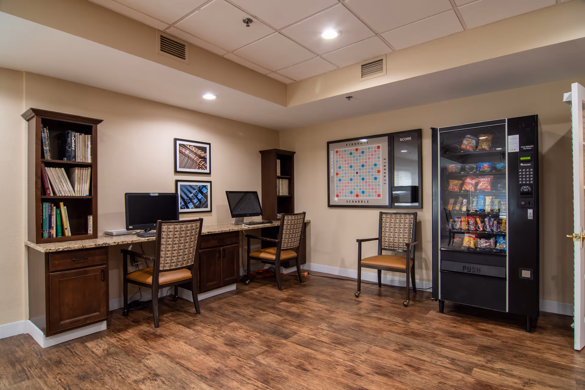 A small community room with two computer workstations on a wooden desk, each with a chair. There are bookshelves on either side of the desk with books. On the wall above the desk are two framed pictures. To the right, there is a framed Scrabble board and a vending machine stocked with snacks. A single chair is placed between the Scrabble board and the vending machine. The room has wood flooring and a beige wall color.