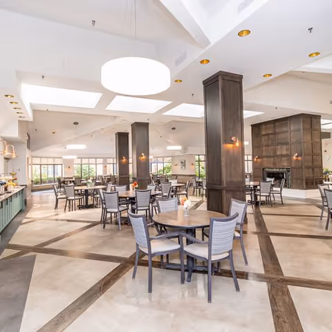 Spacious dining room with multiple round tables and chairs arranged neatly. The room features large windows allowing natural light, modern ceiling lights, and several dark wooden pillars. There is a buffet area on the left side and a fireplace on the right side of the room.