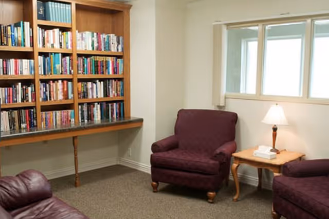 A cozy interior room with a bookshelf filled with books on the left wall, two purple upholstered armchairs, a wooden side table with a lamp and a box of tissues, and a window with white blinds.