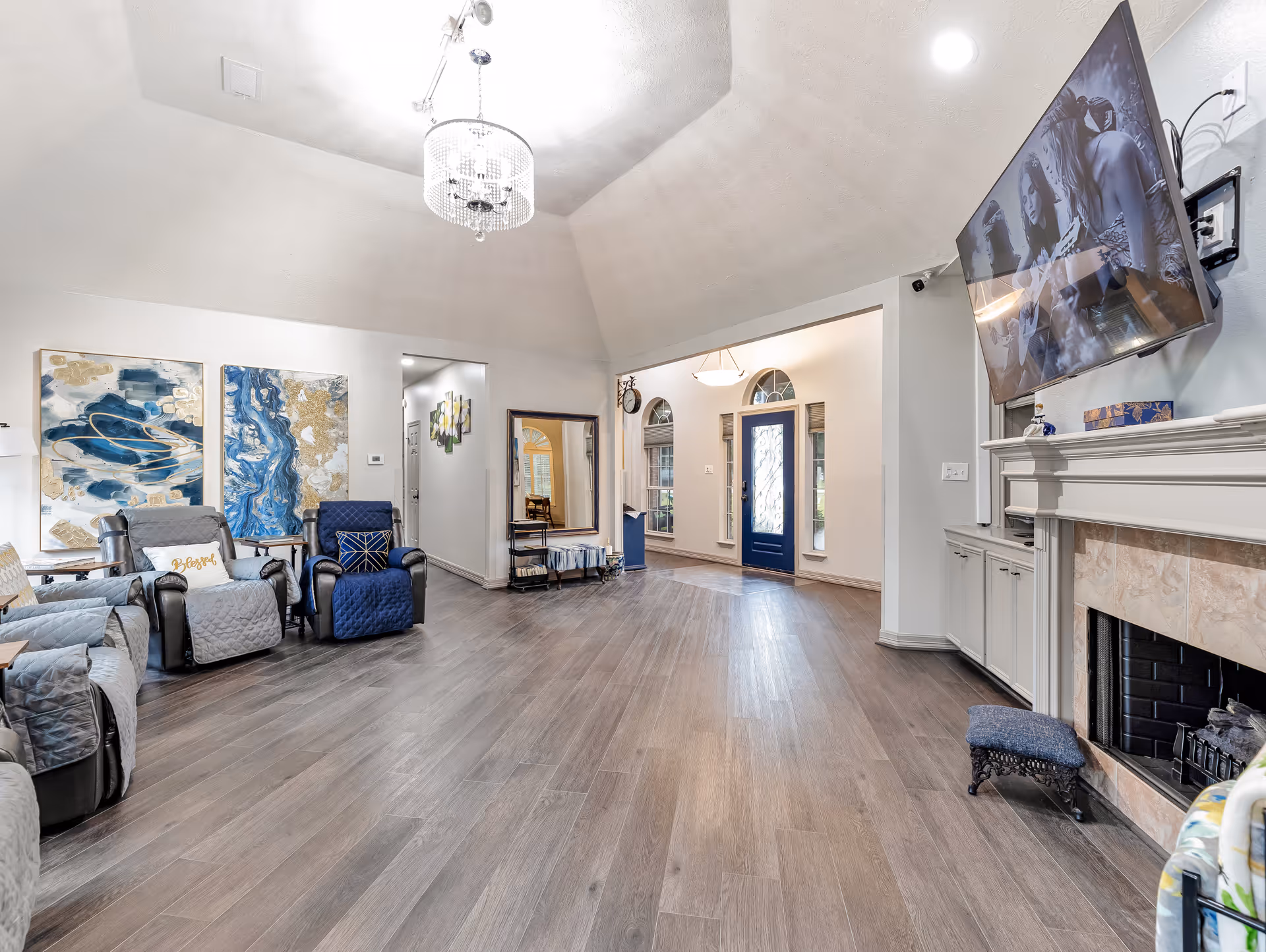 A spacious living room with wooden flooring, several comfortable recliner chairs with cushions, abstract blue and gold wall art, a large mirror, and a fireplace with a mounted flat-screen TV above it. The room has a high ceiling with a chandelier and an entrance door with glass panels and sidelights.