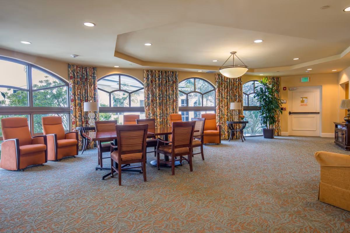 Bright senior living common lounge with a central table and chairs, upholstered armchairs, large arched windows and patterned curtains.
