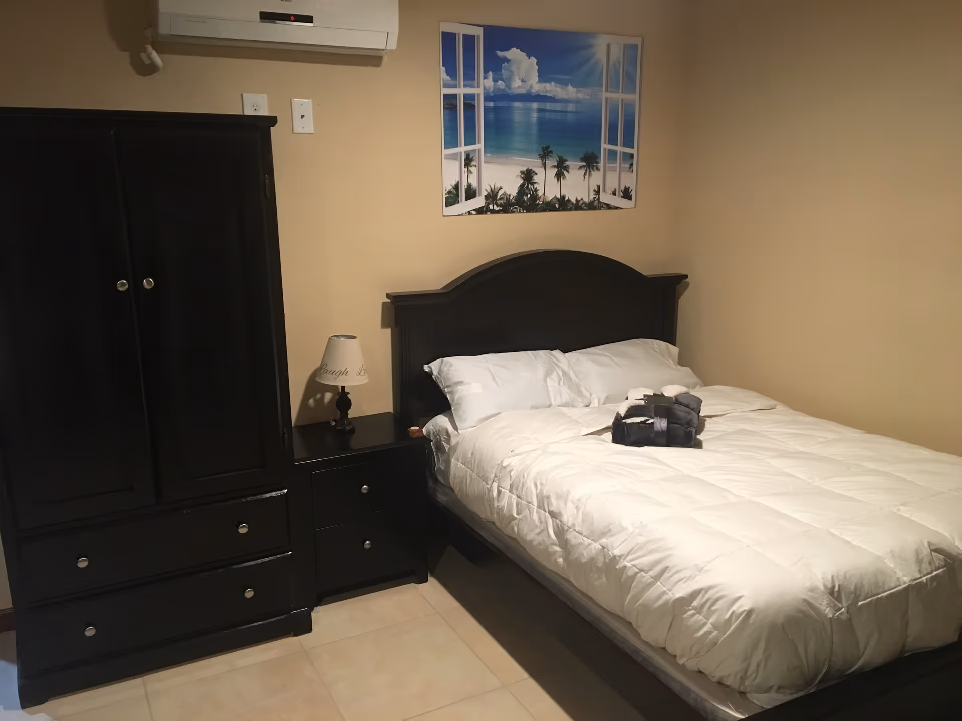 A bedroom with a neatly made bed featuring white bedding and two pillows. On the bed, there are folded towels or robes. Next to the bed is a black nightstand with a small lamp. To the left of the nightstand is a black wardrobe. Above the bed, there is a wall decoration depicting an open window view of a beach with palm trees and blue sky. An air conditioning unit is mounted on the wall above the wardrobe.