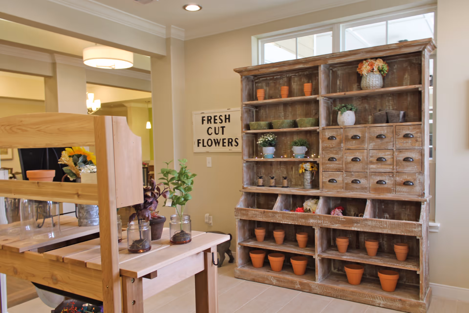 Interior room with a rustic wooden shelving unit holding small flower pots and plants, a wooden table with glass jars containing plants, and a wall sign that reads 'FRESH CUT FLOWERS'. The room is well-lit with beige walls and a window in the background.