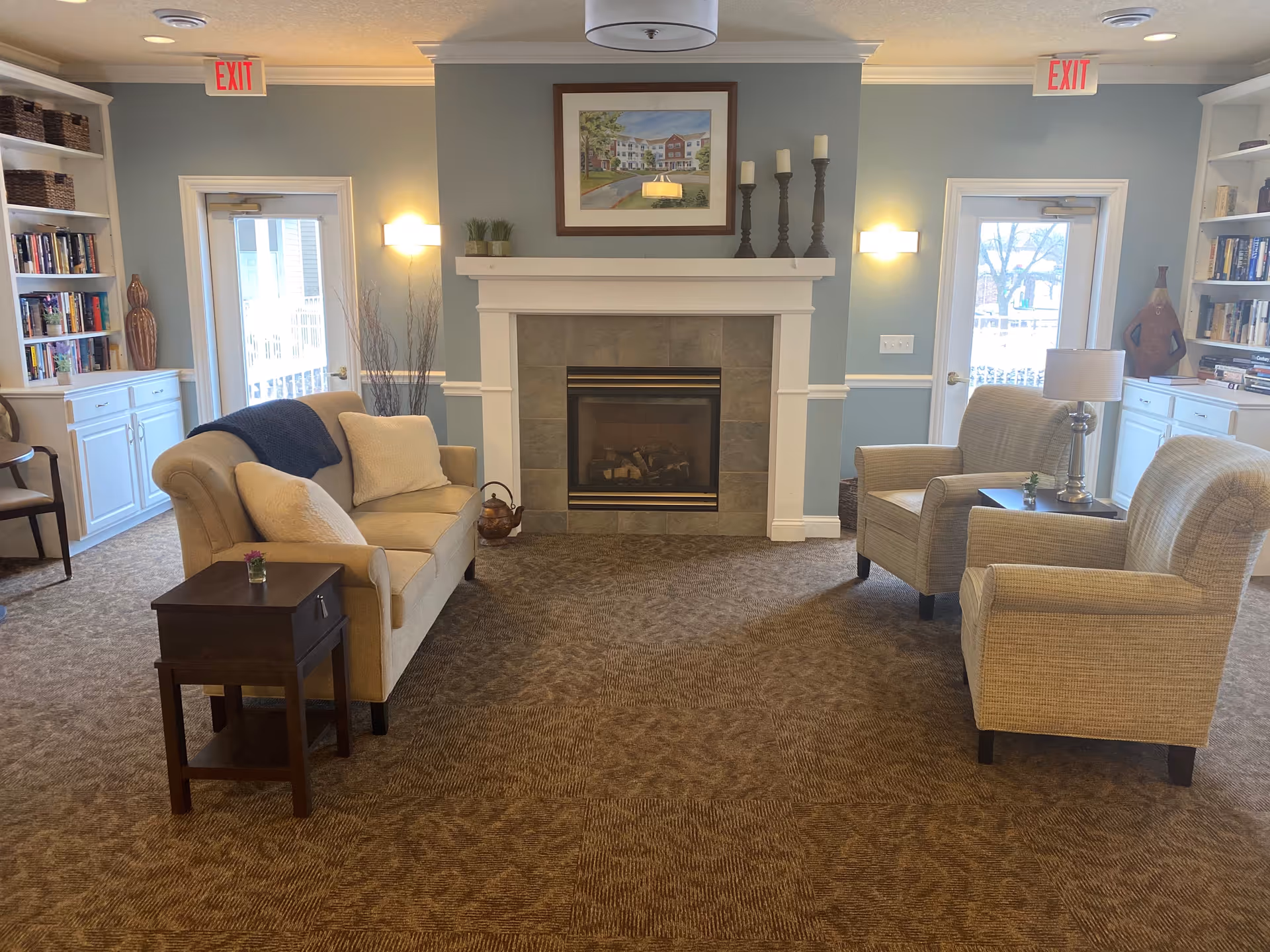 A cozy living room area with a beige sofa and two beige armchairs arranged around a fireplace. The fireplace has a tiled surround and a white mantel with decorative candles and a framed picture above it. There are built-in white bookshelves on either side of the room filled with books and decorative items. Two exit doors with glass panels are visible on either side of the fireplace. The room has soft lighting from wall sconces and a ceiling light fixture.
