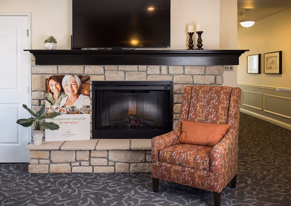 A cozy seating area in a senior living facility featuring a stone fireplace with a black mantel, a mounted flat-screen TV above it, and a floral-patterned armchair with an orange cushion to the right. To the left of the fireplace is a potted plant and a promotional poster showing two smiling elderly women and the text 'Plan a future that's full of life for both of you' with the Vista Springs logo. The area has patterned carpet and a hallway with framed pictures and a ceiling light in the background.