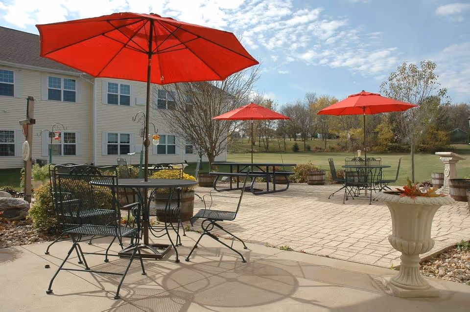 Outdoor patio with metal tables and chairs under red umbrellas in front of a light-colored residential building and lawn.