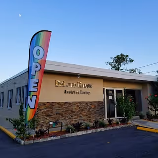 Exterior view of DeLand Manor Assisted Living building during early evening with a clear sky and a visible moon. A rainbow-colored vertical flag with the word 'OPEN' is placed near the entrance. The building has a beige and stone facade with some plants and flowers near the entrance.