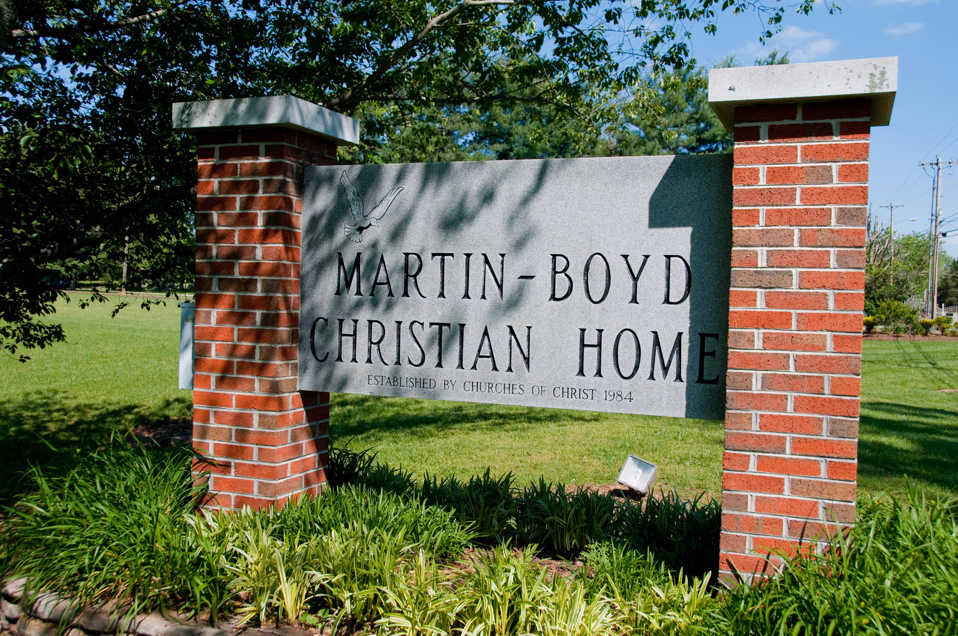 A stone sign supported by two red brick pillars on a grassy lawn. The sign reads 'Martin-Boyd Christian Home Established by Churches of Christ 1984' with an image of a dove above the text. Trees and a clear blue sky are visible in the background.