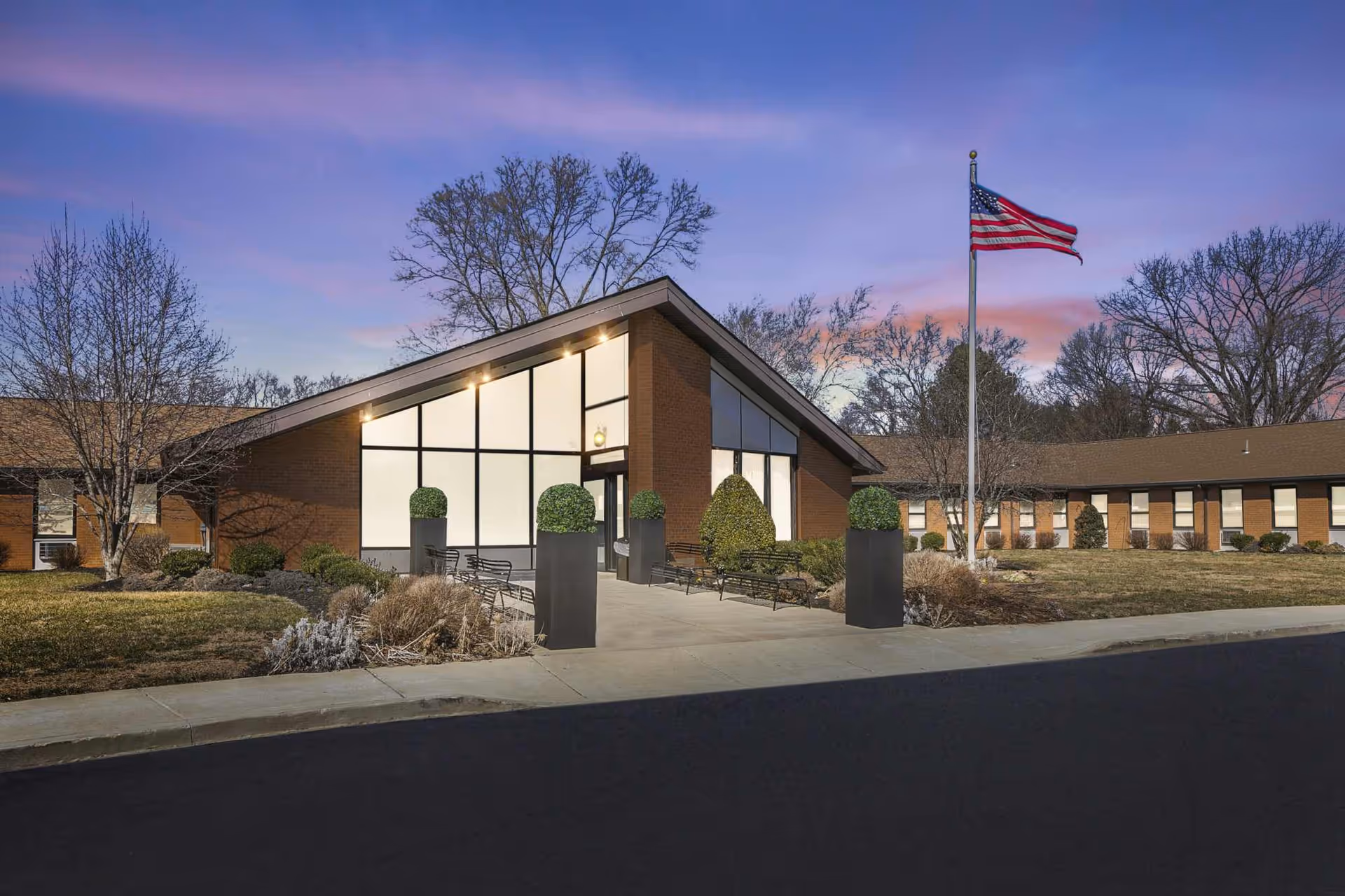 Exterior view of Rosewood Gardens Rehabilitation And Nursing Center building at dusk, featuring a modern entrance with large windows, neatly trimmed bushes in planters, benches along the walkway, an American flag on a flagpole, and leafless trees in the background under a purple and pink sky.