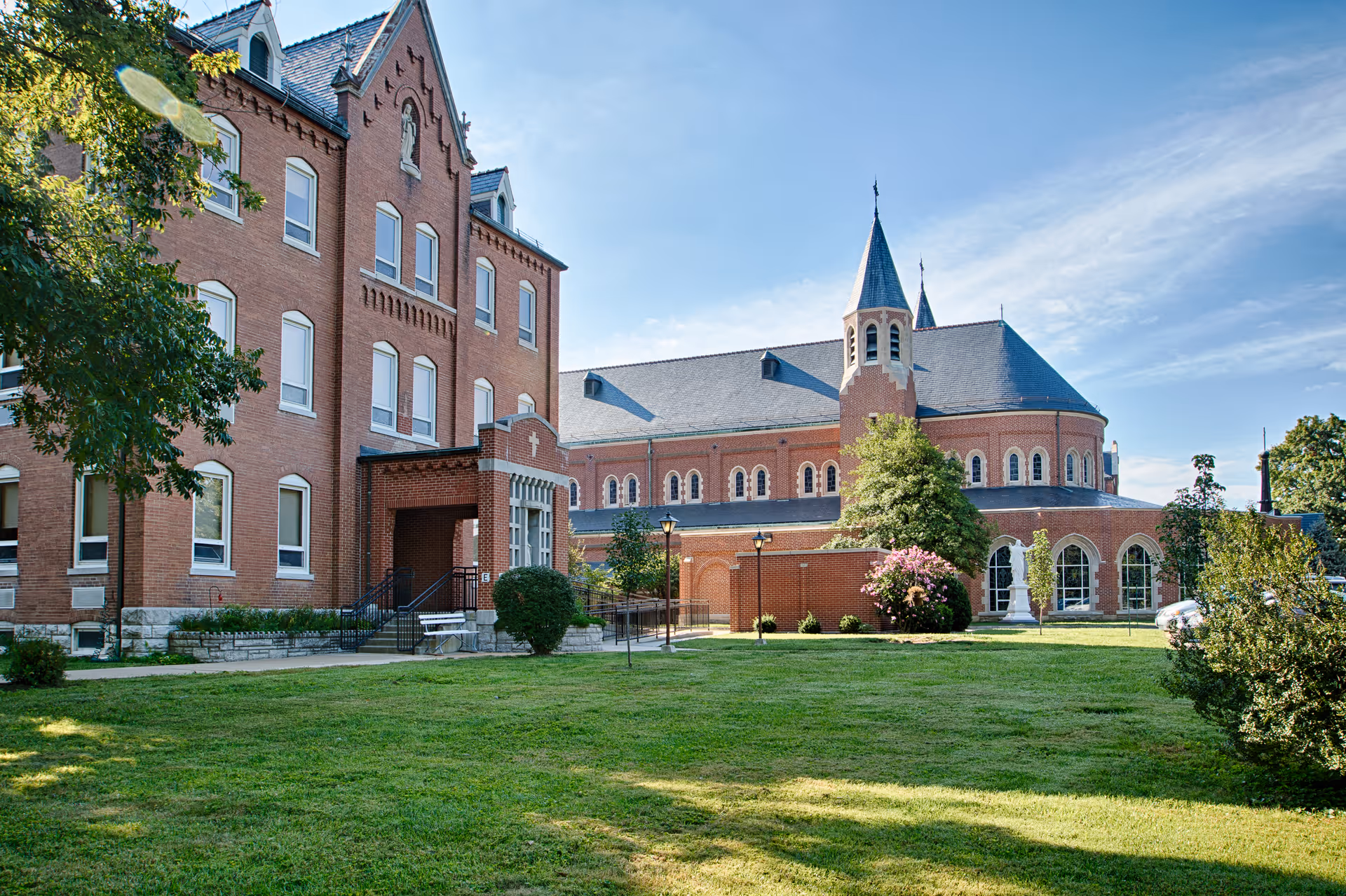 Red-brick multi-story senior living building with an adjoining chapel-like wing and a grassy lawn under a blue sky.