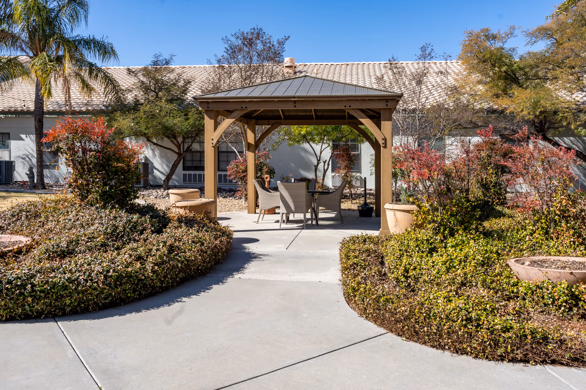 Outdoor patio area with a wooden gazebo covering a table and four chairs, surrounded by landscaped bushes and trees with a building in the background under a clear blue sky.