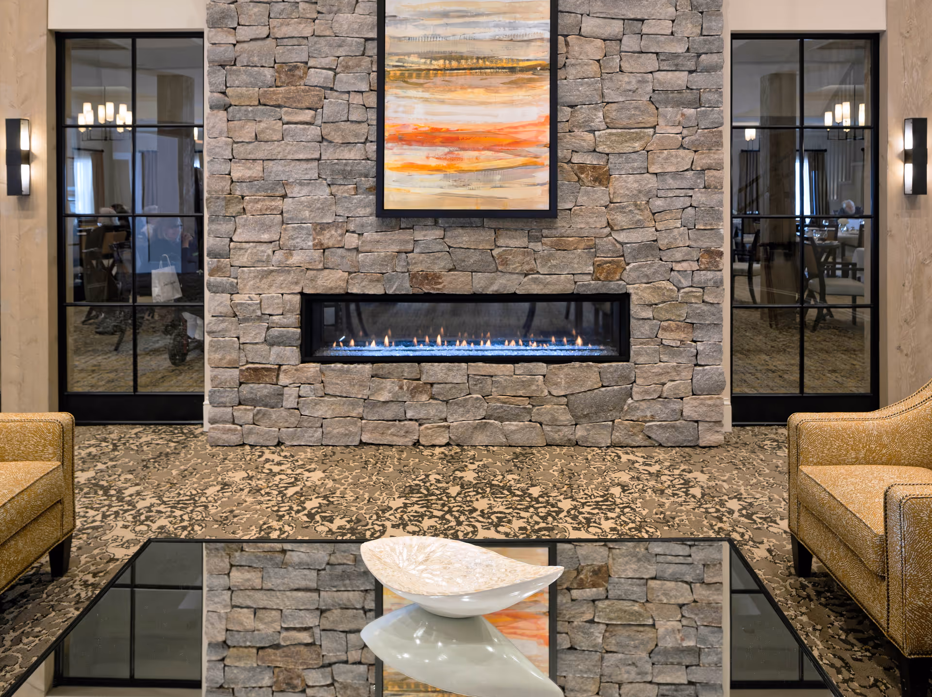 A cozy living room area featuring a stone fireplace with a modern linear gas fire. Above the fireplace hangs an abstract painting with warm tones of orange, yellow, and gray. On either side of the fireplace are black-framed glass doors reflecting the room's interior. Two mustard-yellow upholstered armchairs flank a glass coffee table with a decorative white bowl on top. The floor is covered with a patterned carpet.