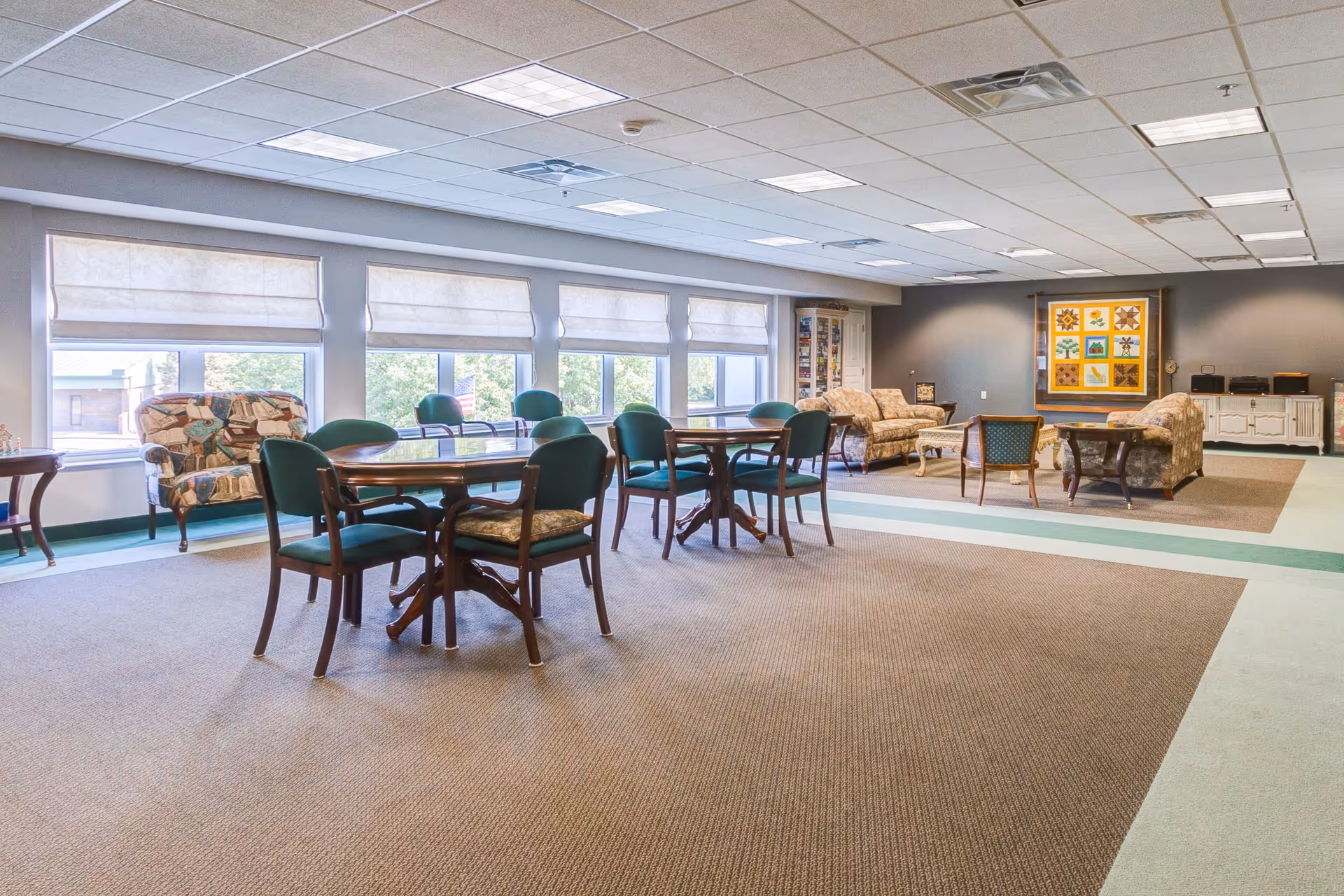 A spacious common area in a senior living facility with multiple seating arrangements including round tables with green cushioned chairs and floral patterned sofas. Large windows with white blinds let in natural light, and a colorful framed quilt hangs on the far wall.