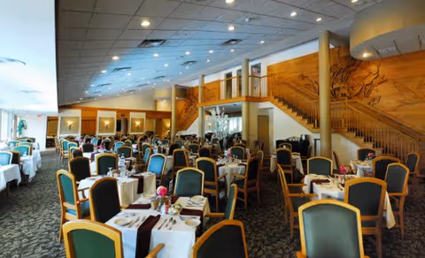 Spacious dining room with numerous tables covered in white tablecloths, set with silverware, napkins, and small flower centerpieces. The room features green and black cushioned chairs, a carpeted floor, and a large wooden staircase with artistic wall carvings in the background.