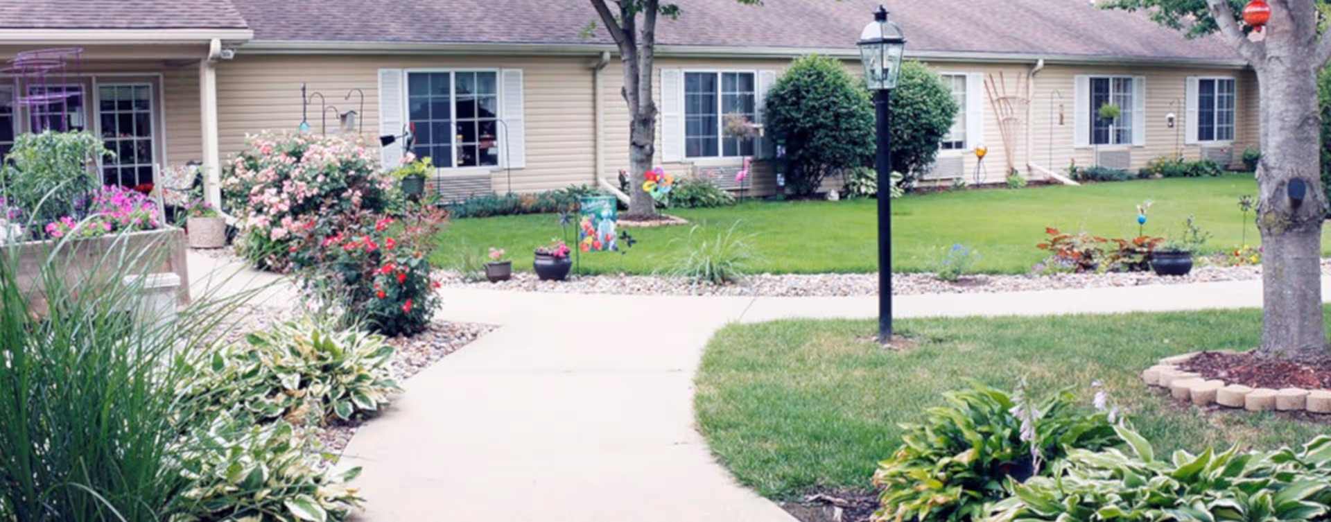 A well-maintained outdoor garden area with a concrete pathway, green grass, various plants, flowers, and shrubs. The background shows a single-story building with beige siding, multiple windows, and a brown roof. There is a black lamp post along the pathway and a tree with a circular stone border around its base.