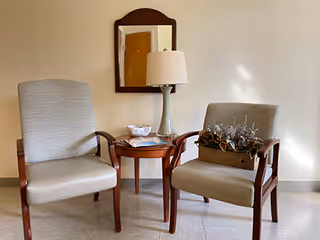 Two cushioned armchairs with wooden frames positioned on either side of a small wooden table. On the table is a lamp with a light green base and a white shade, a small decorative bowl, and some magazines. A rectangular mirror with a wooden frame hangs on the beige wall behind the table and chairs.