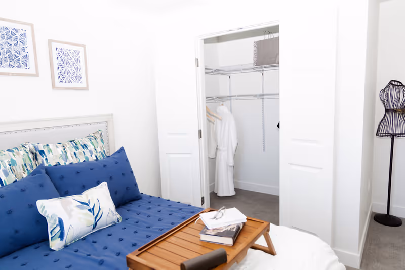A bedroom with a blue bedspread and patterned pillows, a wooden tray with books and glasses on the bed, an open closet with white robes hanging inside, and a decorative dress form standing in the corner.