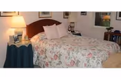 A cozy bedroom with a wooden headboard bed covered in a floral patterned bedspread and two white pillows. There is a round side table with a green tablecloth, a lamp, and framed photos. A window with white curtains and a bookshelf with books are visible in the background.