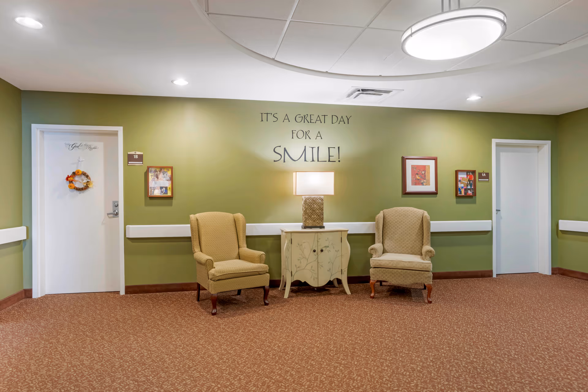 A green-walled hallway seating area with two armchairs, a small painted cabinet and lamp beneath wall lettering that reads "It's a great day for a smile!"