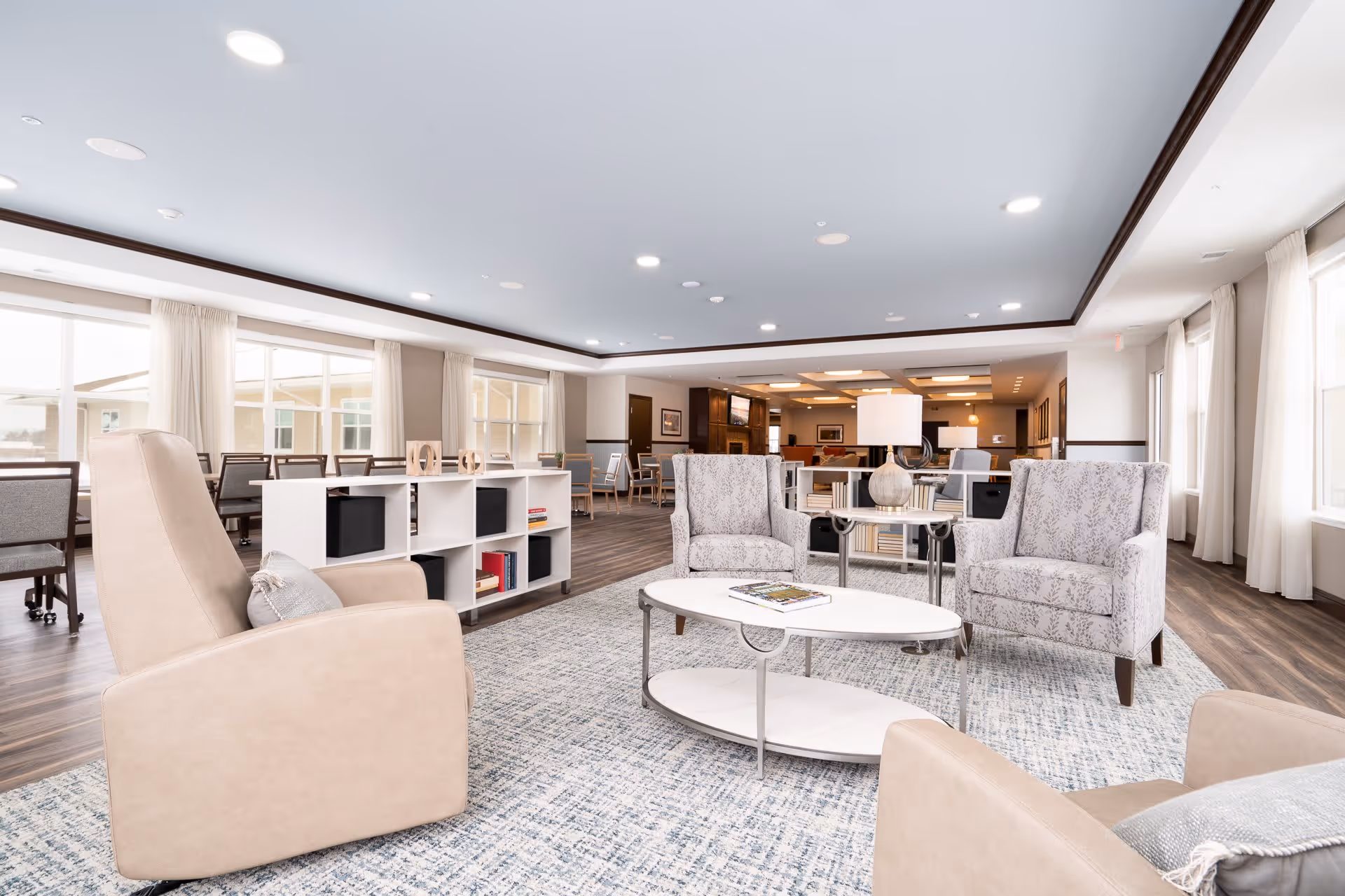 A spacious and well-lit senior living common area with beige and patterned armchairs arranged around a white oval coffee table on a blue and white patterned rug. Behind the seating area is a white cubed shelving unit with books and decorative items. Large windows with cream curtains allow natural light to fill the room. In the background, there are more chairs and tables, and a television mounted on the wall.