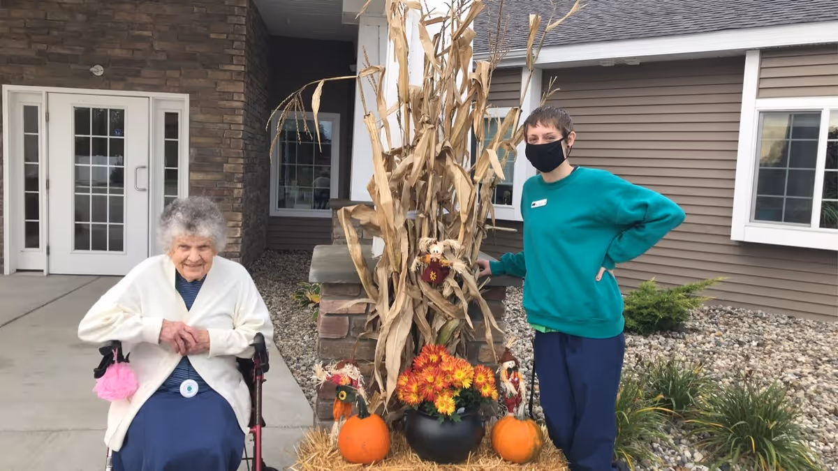 An elderly woman sitting in a wheelchair outside a building next to a younger person wearing a green sweatshirt and a black face mask. They are standing near a fall-themed decoration with dried corn stalks, orange pumpkins, and a pot of yellow and orange flowers. The building has brown siding, white-framed windows, and a stone pillar.