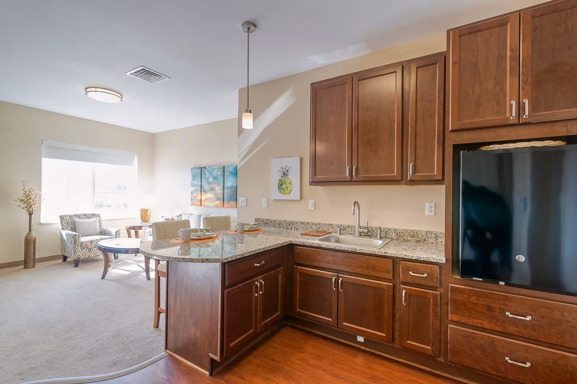 A bright and clean kitchen area with dark wooden cabinets, granite countertops, and a black refrigerator. The kitchen opens into a living room with a patterned armchair, a small round coffee table, and a window letting in natural light. The walls are light-colored and decorated with simple artwork.