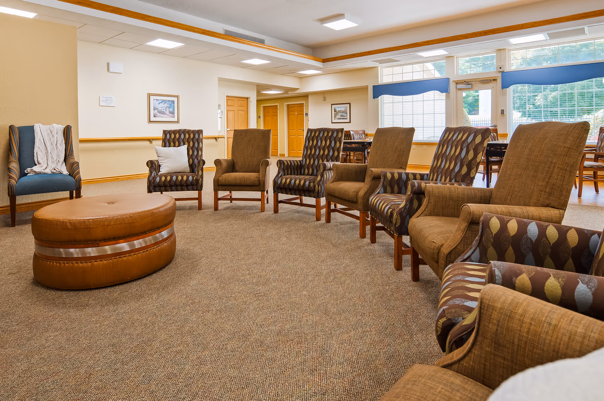 A spacious, well-lit common area with a circle of upholstered armchairs and a large round ottoman in the center. The room has beige walls, carpeted flooring, large windows with blinds, and wooden trim. There are framed pictures on the walls and a dining area visible in the background.