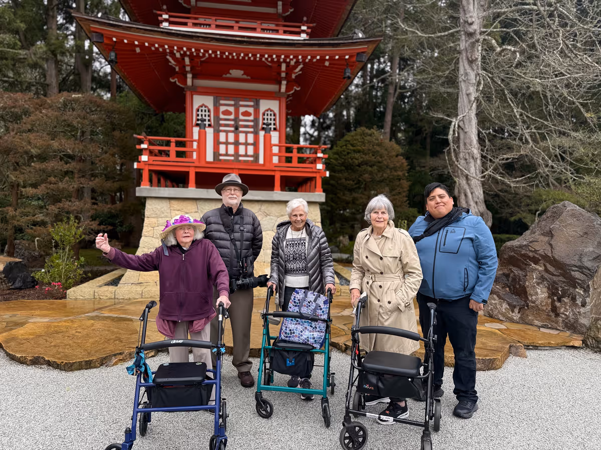 Five people standing outdoors in front of a red and white pagoda-style structure surrounded by trees and rocks. Three elderly women using walkers are accompanied by an elderly man wearing a hat and a younger man in a blue jacket. The ground is covered with gravel and stone paving.