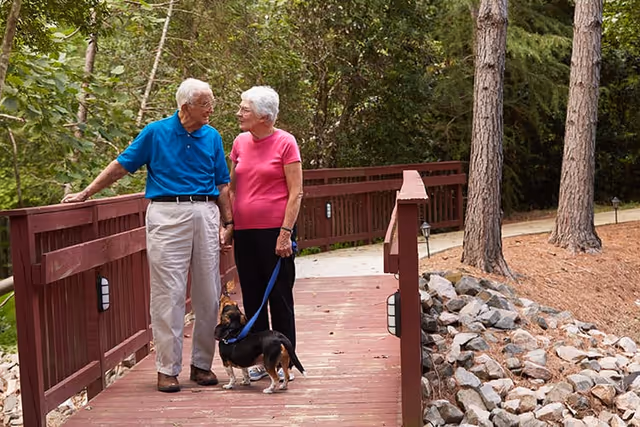 An elderly couple holding hands and standing on a wooden bridge in a wooded outdoor area. The woman is holding a leash attached to a small dog. Both are smiling and looking at each other.