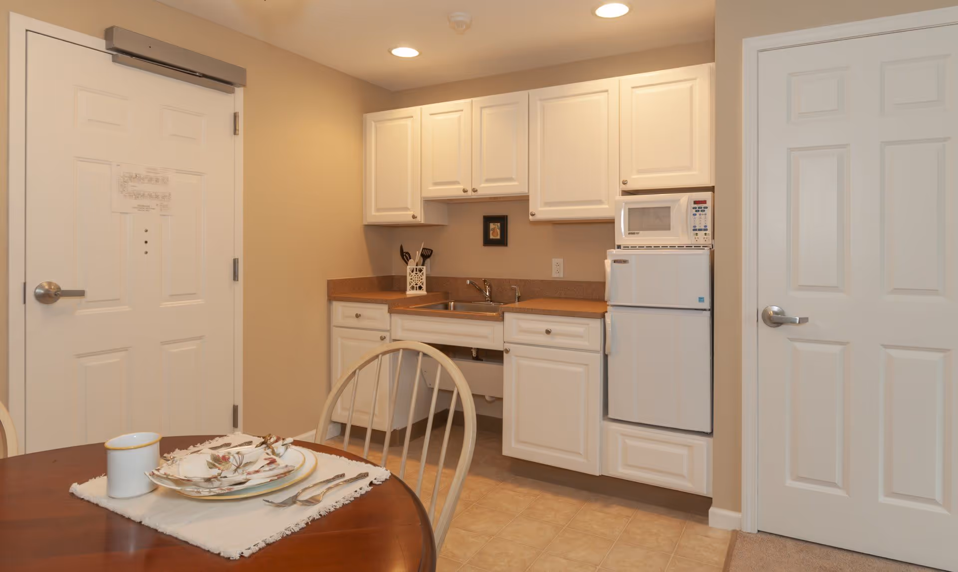 Small kitchenette with white cabinets, a sink, mini-fridge and microwave, and a dining table with place settings in the foreground.