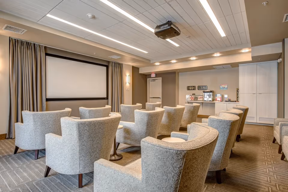 A cozy movie theater room with multiple rows of comfortable beige armchairs facing a large blank projector screen. The room has a ceiling-mounted projector, soft lighting, and a small concession area with popcorn and snacks at the back.