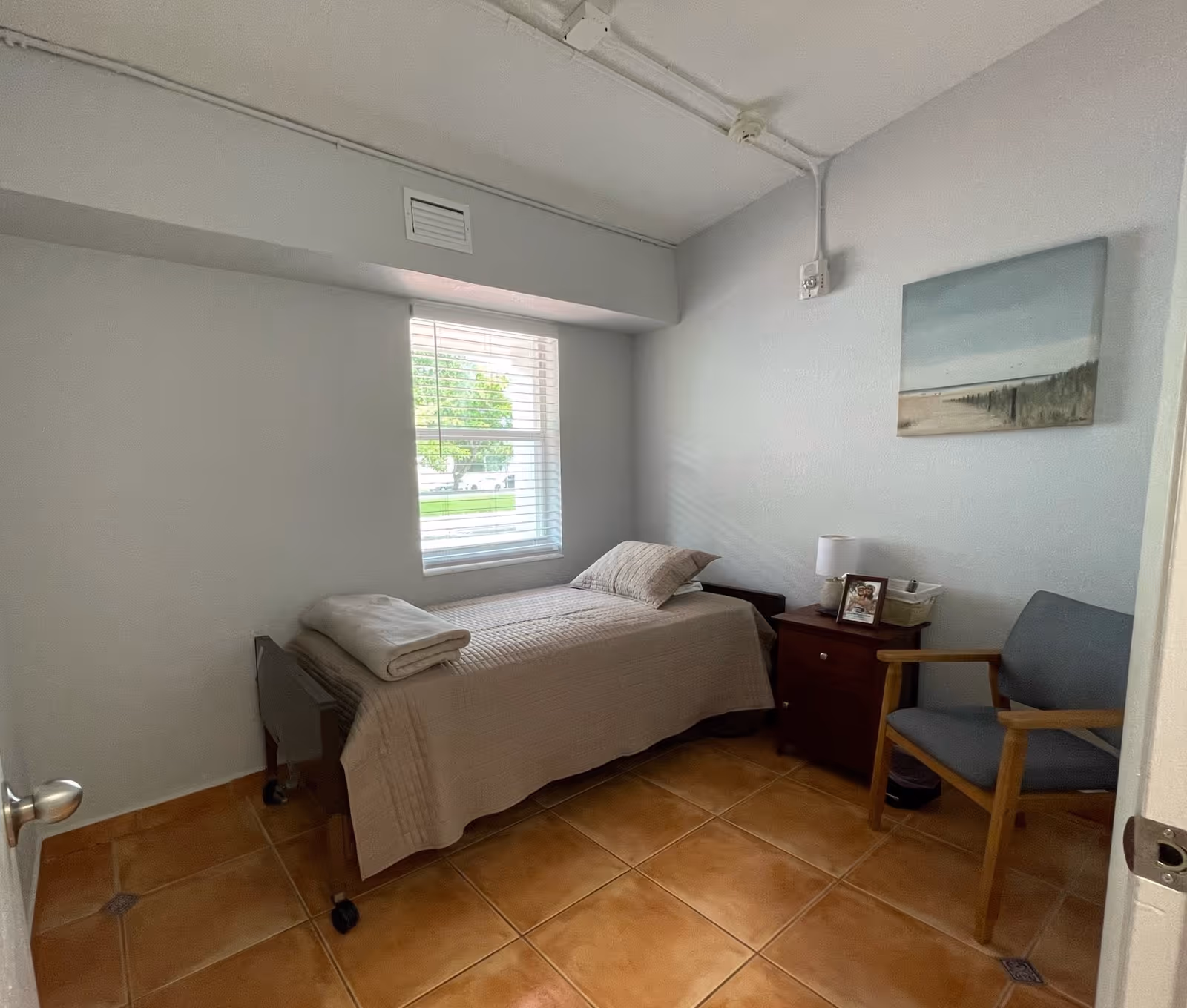Small furnished bedroom with a single bed, bedside table with lamp and framed photo, a chair, and a window.