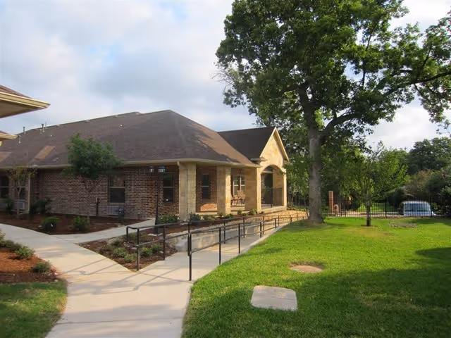 Single-story brick building with a sloped roof, surrounded by a well-maintained lawn and trees. A concrete pathway with a handrail leads to the entrance of the building under a covered porch. The sky is partly cloudy.