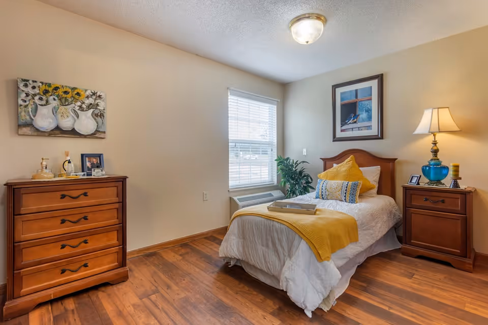 Cozy single bedroom with a bed dressed in white and yellow accents, wooden nightstand and dresser, window, and wall art.