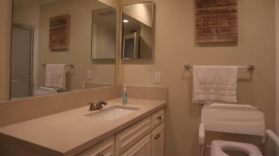 A bathroom with a beige countertop and sink, a large mirror above the sink, a towel rack with a white towel, a white shower chair, and a wooden wall decoration with an inspirational quote.