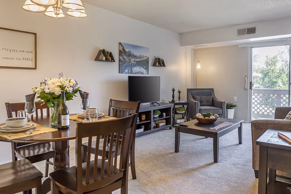 A cozy living and dining area in a senior living facility. The dining area features a round wooden table set with plates, bowls, wine glasses, and a bottle of wine, along with a vase of white and purple flowers. The living area includes a TV on a stand, a coffee table with decorative items, an armchair, and a sliding glass door leading to an outdoor balcony. The walls are decorated with a framed quote and a landscape photograph.