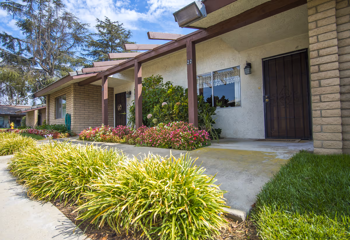 Exterior view of a single-story residential building with a covered walkway supported by wooden beams. The building has beige brick and stucco walls, a black metal security door, and a window with lace curtains. There are well-maintained flower beds with green shrubs and pink flowers along the walkway, and a green lawn beside the concrete path. Trees and a blue sky with some clouds are visible in the background.