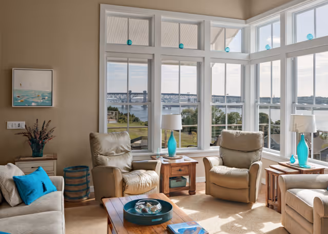 A bright living room with large windows offering a scenic view of a river and a bridge. The room features two beige leather armchairs, a beige sofa with blue cushions, wooden side tables with turquoise lamps, a coffee table with decorative items, and a painting on the wall.